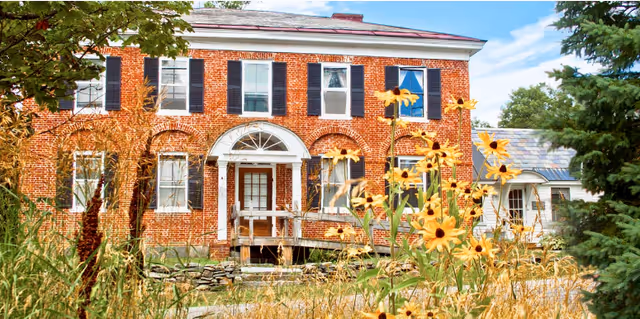 A large two-story brick building with white trim and black shutters, featuring a central entrance with a white arched doorway. The foreground shows tall grasses and yellow wildflowers, with trees on either side under a partly cloudy sky.