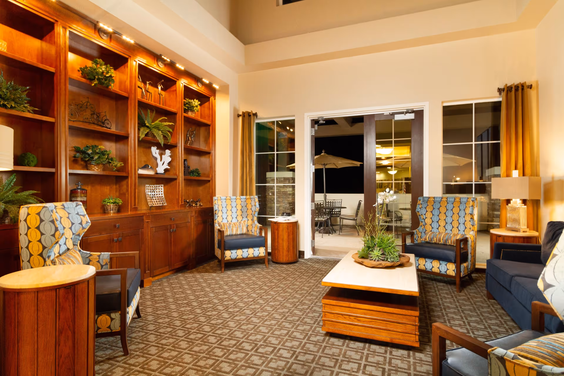A cozy living room area with patterned armchairs, a wooden coffee table with a plant centerpiece, a dark blue sofa, and a large wooden built-in bookshelf decorated with plants and ornaments. Glass doors open to an outdoor patio with a table and umbrella visible.