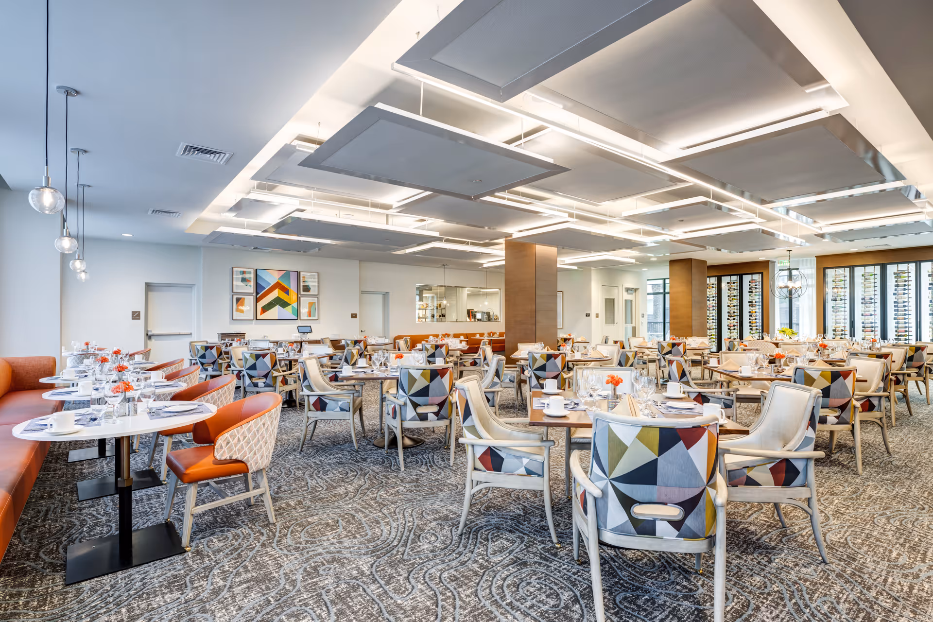 A spacious, modern dining room with multiple tables set for meals. The room features patterned carpet, colorful geometric upholstered chairs, orange cushioned bench seating along one wall, and contemporary ceiling light fixtures. There are decorative artworks on the walls and large windows with wine racks in the background.