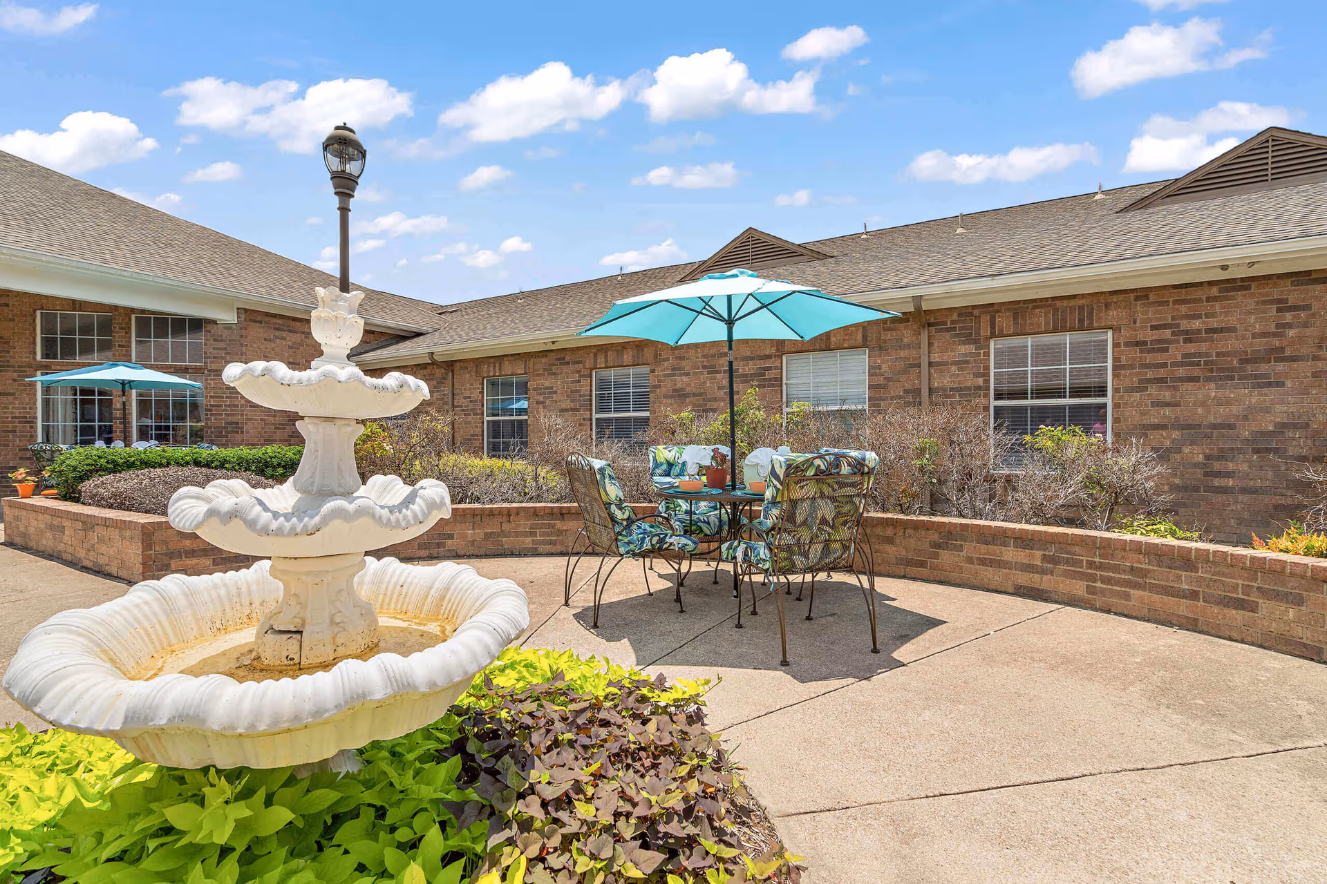 Outdoor patio area at Morada Cedar Hill featuring a white tiered fountain surrounded by green and purple plants, a round metal table with four cushioned chairs under a turquoise umbrella, and a brick building with windows in the background under a partly cloudy blue sky.