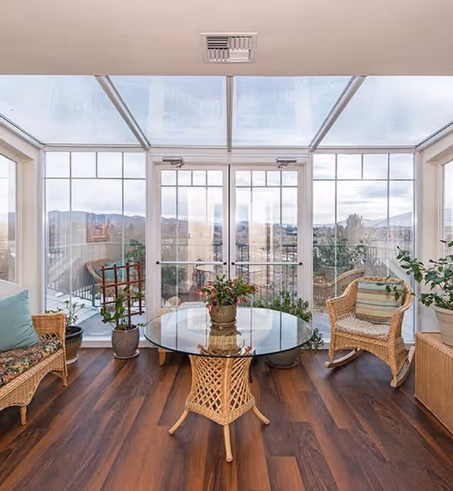 A bright sunroom with large glass windows and a glass ceiling, featuring wicker furniture including a cushioned sofa, a chair with a striped pillow, and a glass-top table with a plant centerpiece. Several potted plants are placed around the room, and there is a scenic view of mountains and trees outside.