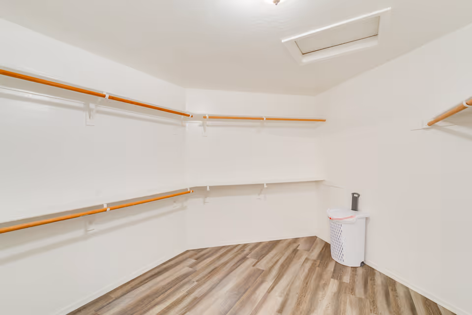 Empty walk-in closet with white walls, wooden hanging rods on three sides, a white laundry basket with a trash bag in the corner, and wood-patterned flooring.