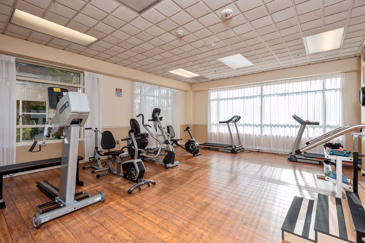 A bright exercise room with wooden flooring and large windows covered with sheer white curtains. The room contains various fitness equipment including treadmills, stationary bikes, and other exercise machines. The ceiling has a grid pattern with recessed lighting.