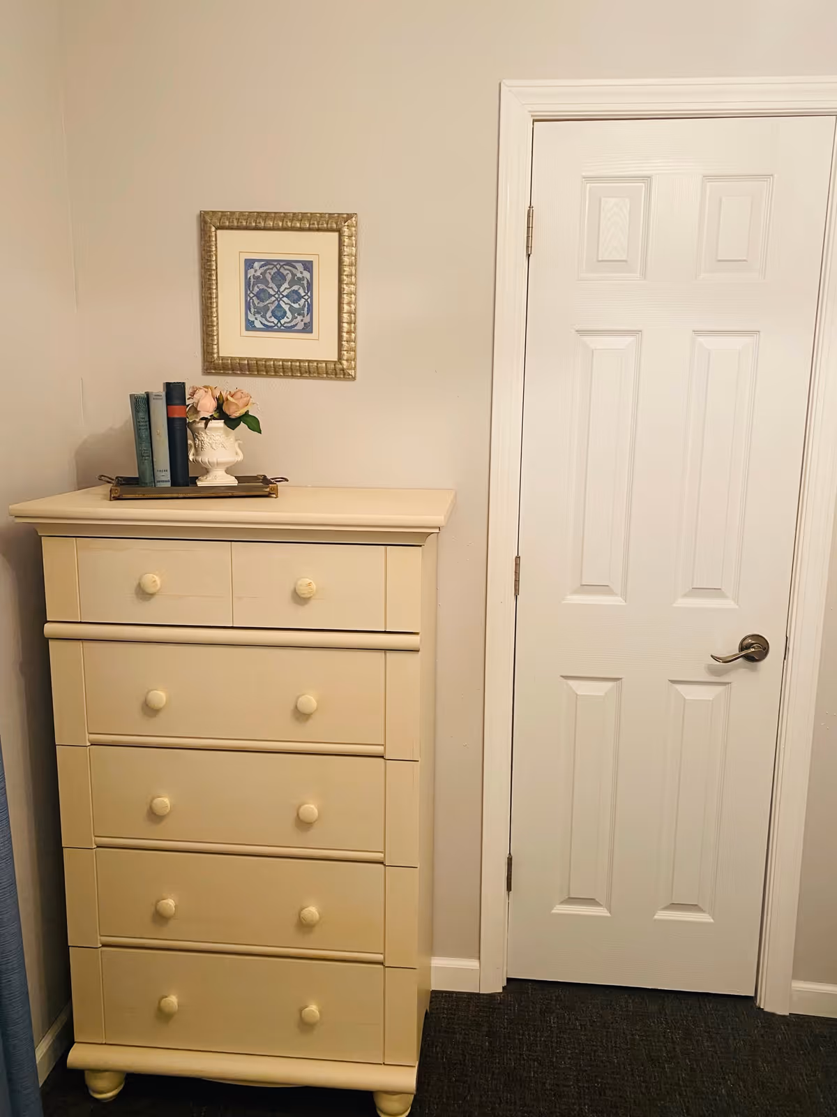 A cream-colored chest of drawers with five drawers stands next to a closed white door with a silver handle. On top of the chest is a tray holding three books and a white vase with pink flowers. Above the chest hangs a framed piece of artwork with a blue and white pattern. The walls are light beige and the floor is covered with dark carpet.