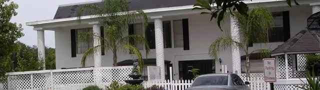 Two-story white building with tall columns, palm trees, a white picket fence, and a car parked in front.