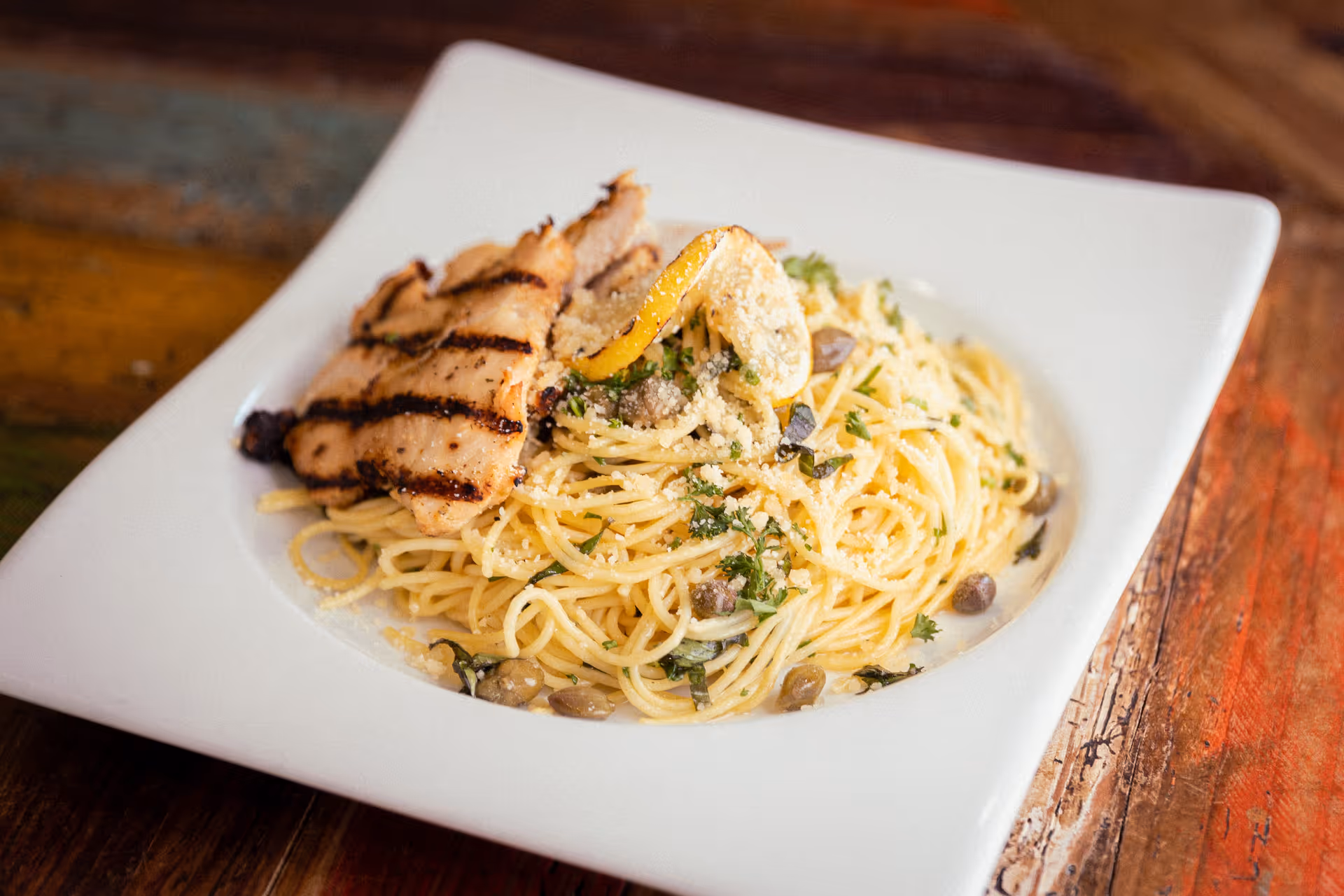 A white square plate with a serving of spaghetti pasta garnished with herbs, capers, grated cheese, and a slice of lemon, topped with two pieces of grilled chicken breast with grill marks, placed on a rustic wooden table.
