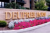 A brick sign with large gold letters spelling 'DEUPREE HOUSE' surrounded by landscaping including pink flowers and green shrubs, with part of a building and trees visible in the background.