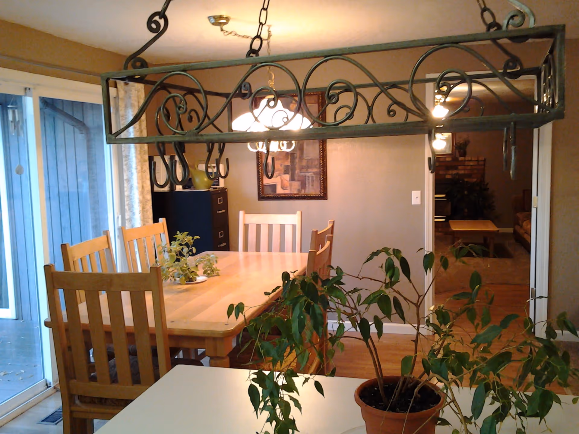 A dining room with a wooden table and six chairs. A decorative metal hanging rack with hooks is suspended above the table. There are two potted plants in the foreground, one on a white surface and another on the dining table. A sliding glass door with curtains is on the left side, and a doorway leads to a living room area with a couch, coffee table, and a brick fireplace in the background.