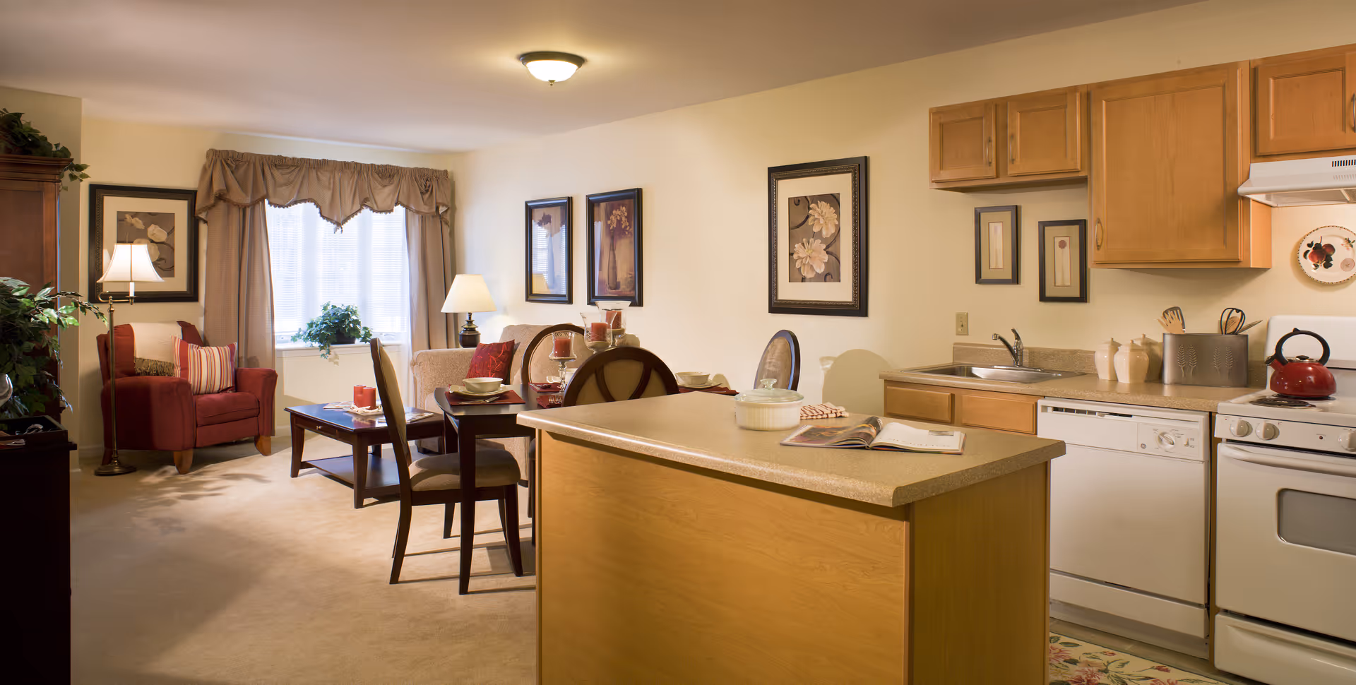 A cozy senior living apartment interior featuring a small kitchen with wooden cabinets, a white stove, dishwasher, and a kitchen island. Adjacent to the kitchen is a dining area with a table set for four, and a living area with a red armchair, coffee table, and beige sofa. The room is warmly lit with lamps and natural light from a window with curtains.