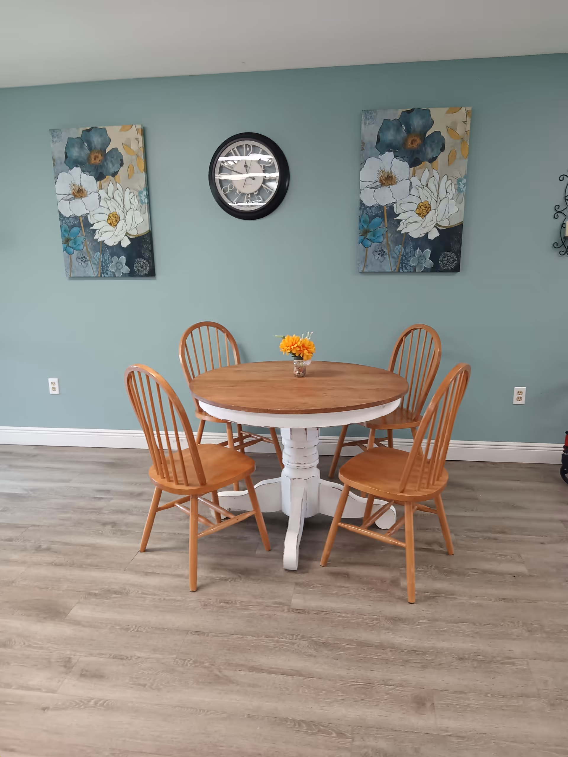 A round wooden dining table with a white base surrounded by four wooden chairs with vertical slats. A small vase with yellow flowers is placed on the table. The background features a light blue wall with two floral paintings and a round wall clock in the center.