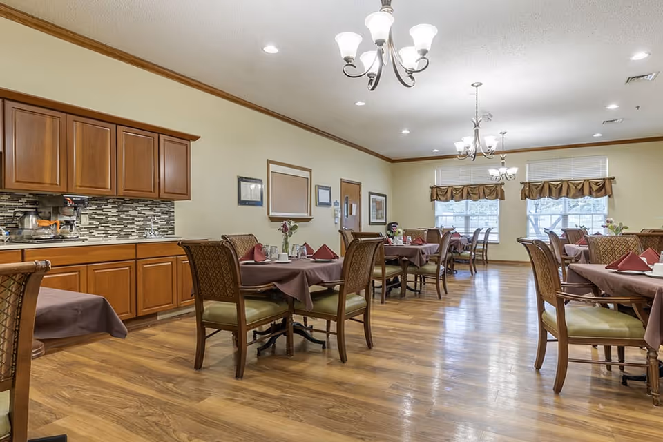 A dining room in a senior living facility with multiple tables covered in brown tablecloths, each set with napkins, cups, and small flower arrangements. The room has wooden flooring, beige walls, wooden cabinets along one side, and large windows with brown valances letting in natural light. Ceiling lights with multiple bulbs hang above the tables.