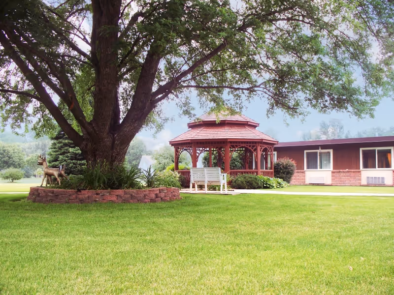 A large tree with a circular brick planter at its base, a white bench, and a red wooden gazebo in a well-maintained grassy outdoor area. In the background, there is a single-story building with brown and brick exterior walls and windows.