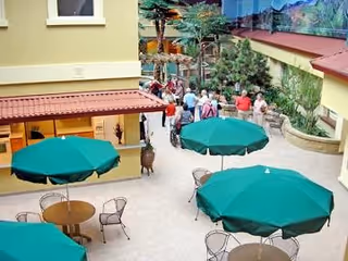 Open courtyard with green patio umbrellas, round tables and chairs, surrounded by building walls and a group of people near a mural.