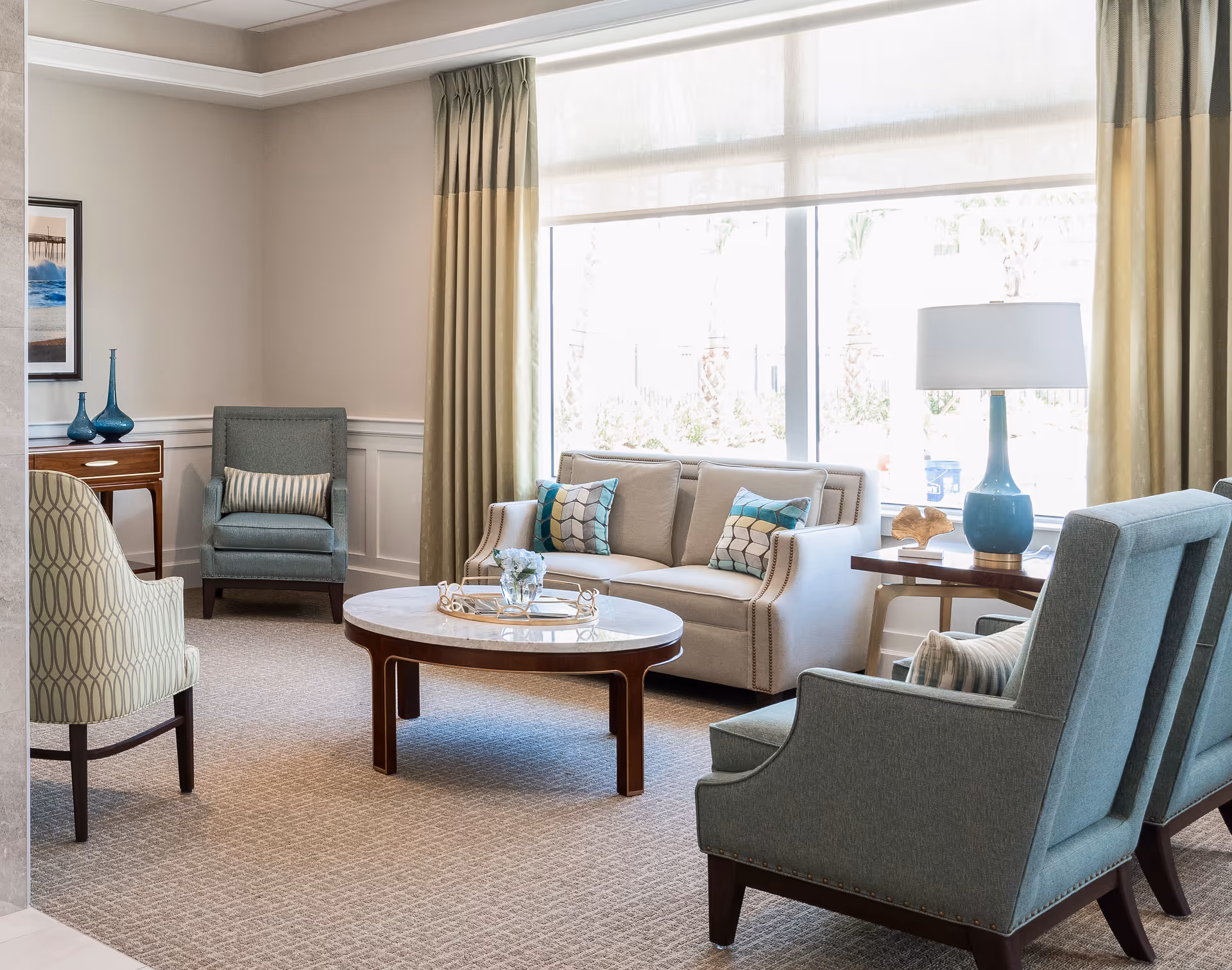 A bright and cozy living room with a beige sofa adorned with patterned cushions, two blue armchairs, and a patterned beige chair around a round wooden coffee table. A side table with a blue lamp and decorative items is next to the sofa. Large windows with beige curtains allow natural light to fill the room.