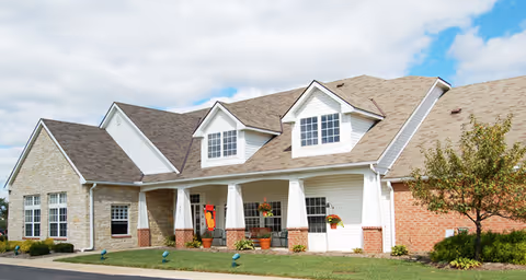 Exterior view of a single-story residential building with a combination of brick and stone facade, white siding, multiple windows, and a front porch with potted plants and hanging flower baskets under a partly cloudy sky.