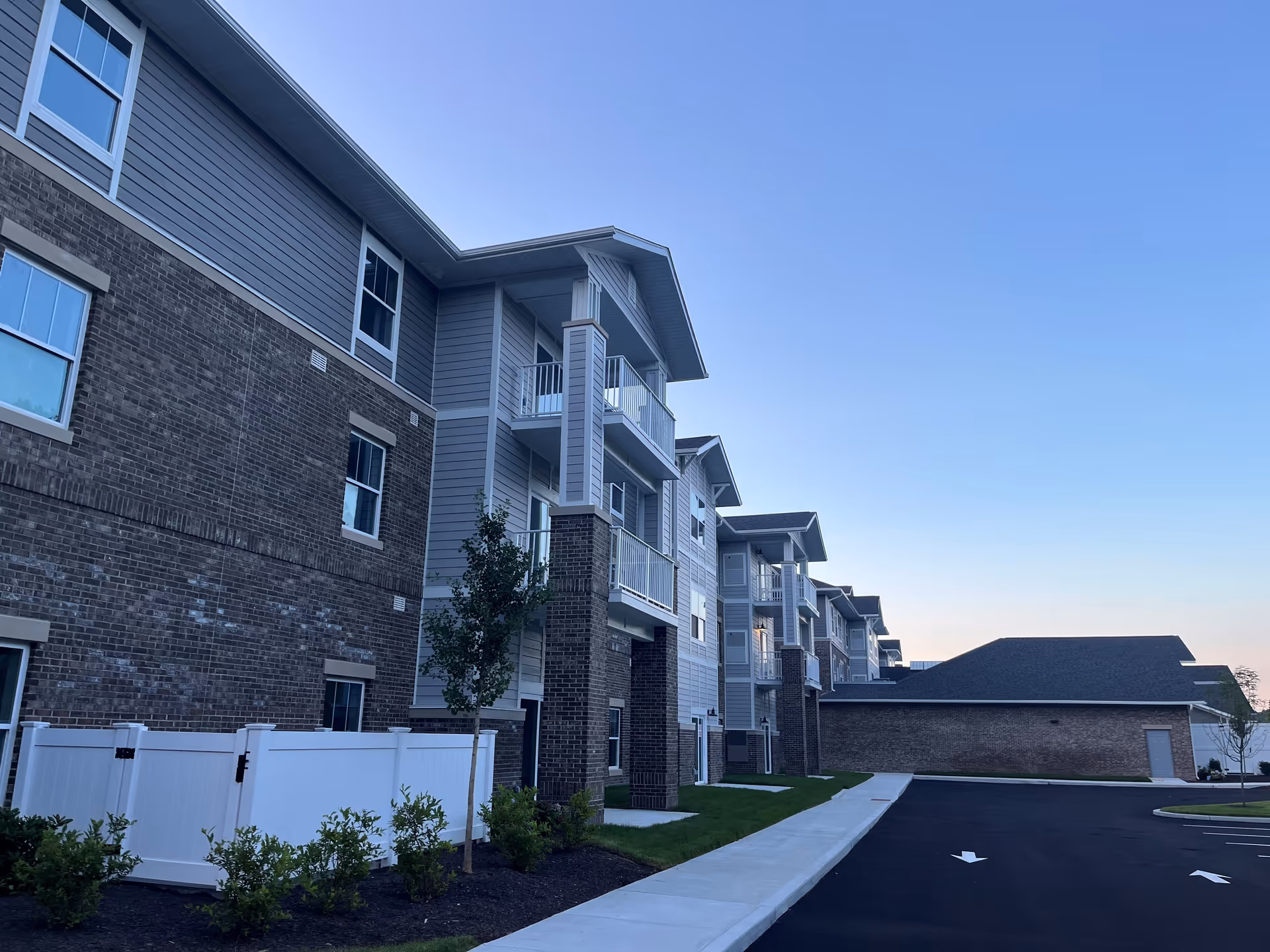 Exterior view of a multi-story brick-and-siding retirement apartment building with balconies, landscaping, and a parking area under a clear sky.