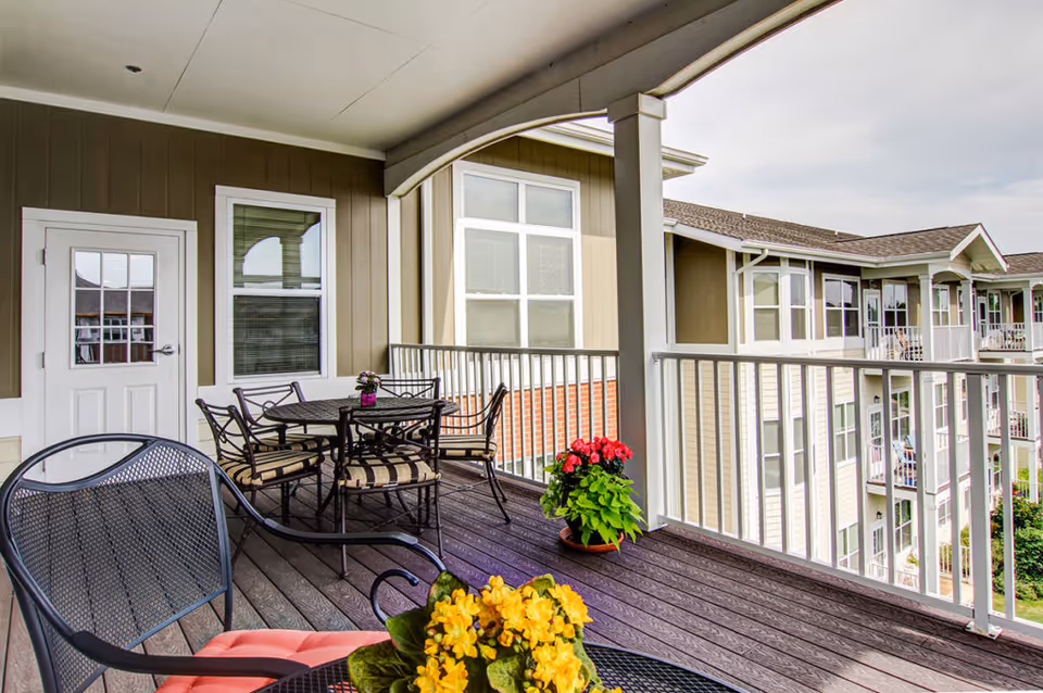 Covered balcony with a table and chairs, potted flowers, and a view of neighboring apartment balconies.