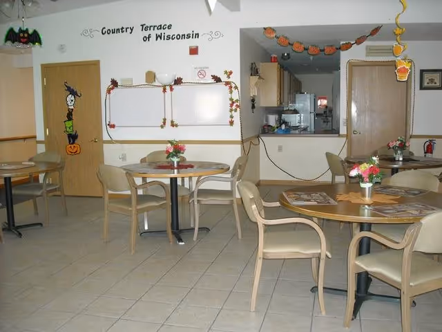 Interior view of a dining area at Country Terrace of Wisconsin with several round tables and beige chairs. The tables have small flower arrangements and placemats. The room is decorated with Halloween-themed garlands and hanging decorations. There is a kitchen pass-through window in the background and a whiteboard on the wall with the facility name above it.