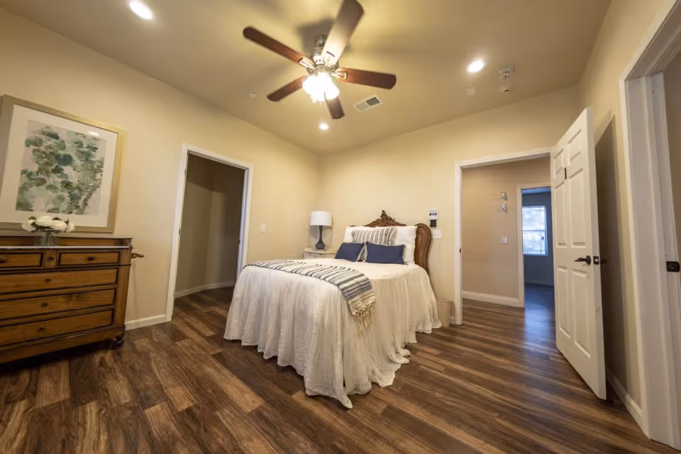 A bedroom with a wooden floor, a bed with white bedding and blue pillows, a wooden dresser with a framed artwork above it, a ceiling fan with lights, and two open doorways leading to other rooms.