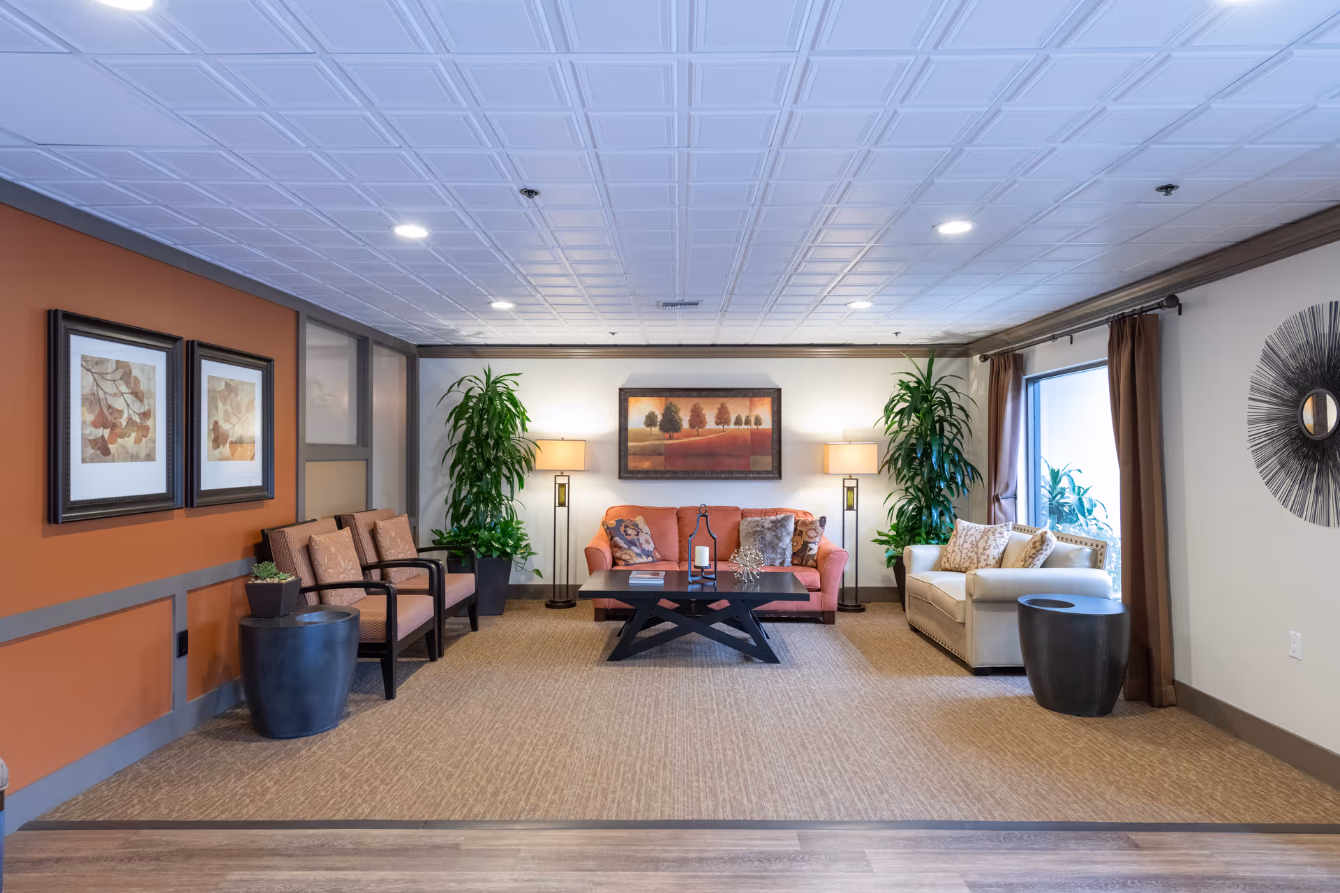 A cozy living room area with a coral sofa and a white sofa, both adorned with decorative pillows. A black coffee table sits in the center with a candle and decor items. Two tall floor lamps flank the coral sofa, and two large green plants are placed behind the sofas. The walls are decorated with framed artwork and a circular wall mirror. The room has a beige carpet and a white ceiling with recessed lighting.