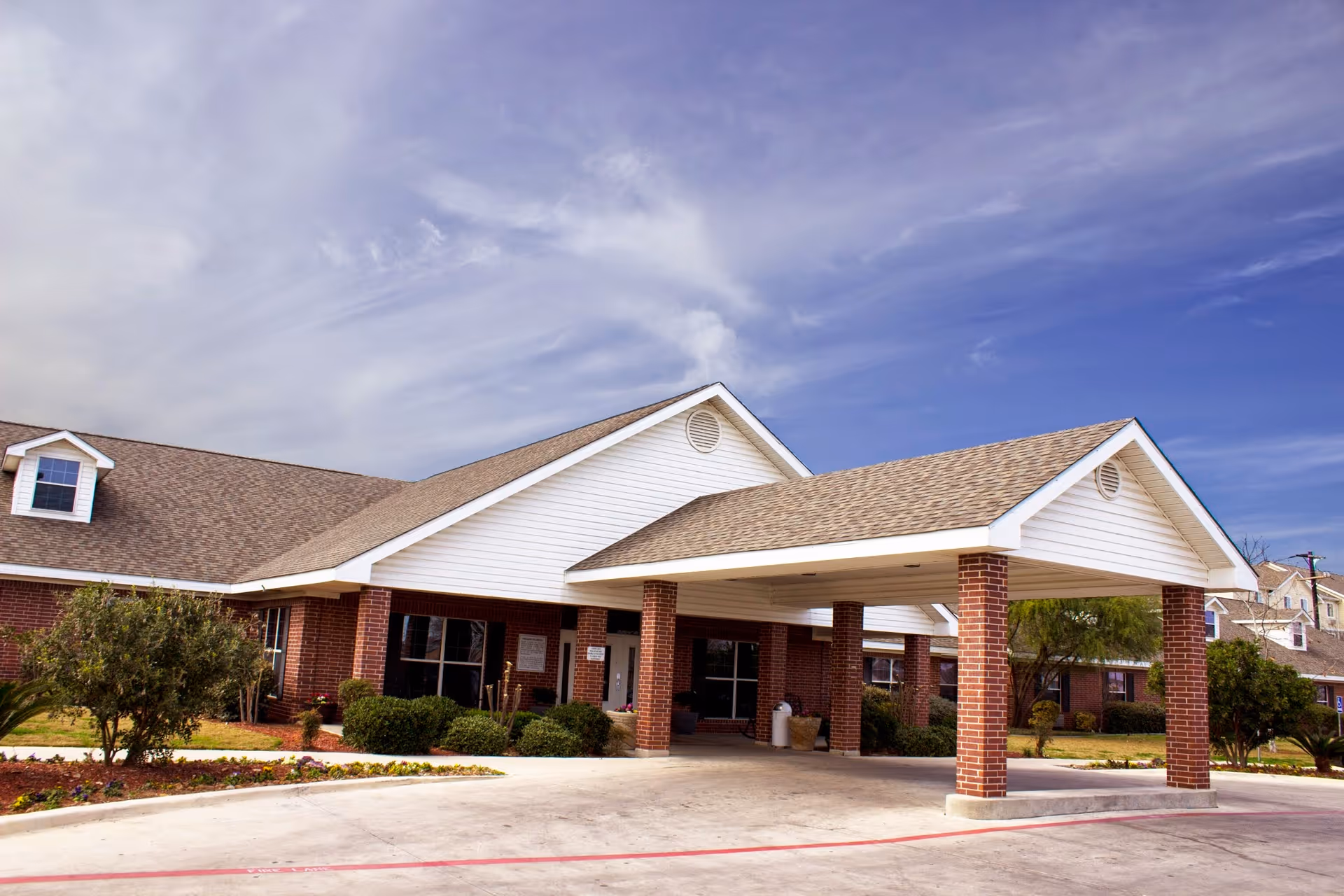 Front exterior view of a single-story brick building with a covered entrance supported by brick columns, surrounded by landscaping and under a partly cloudy sky.