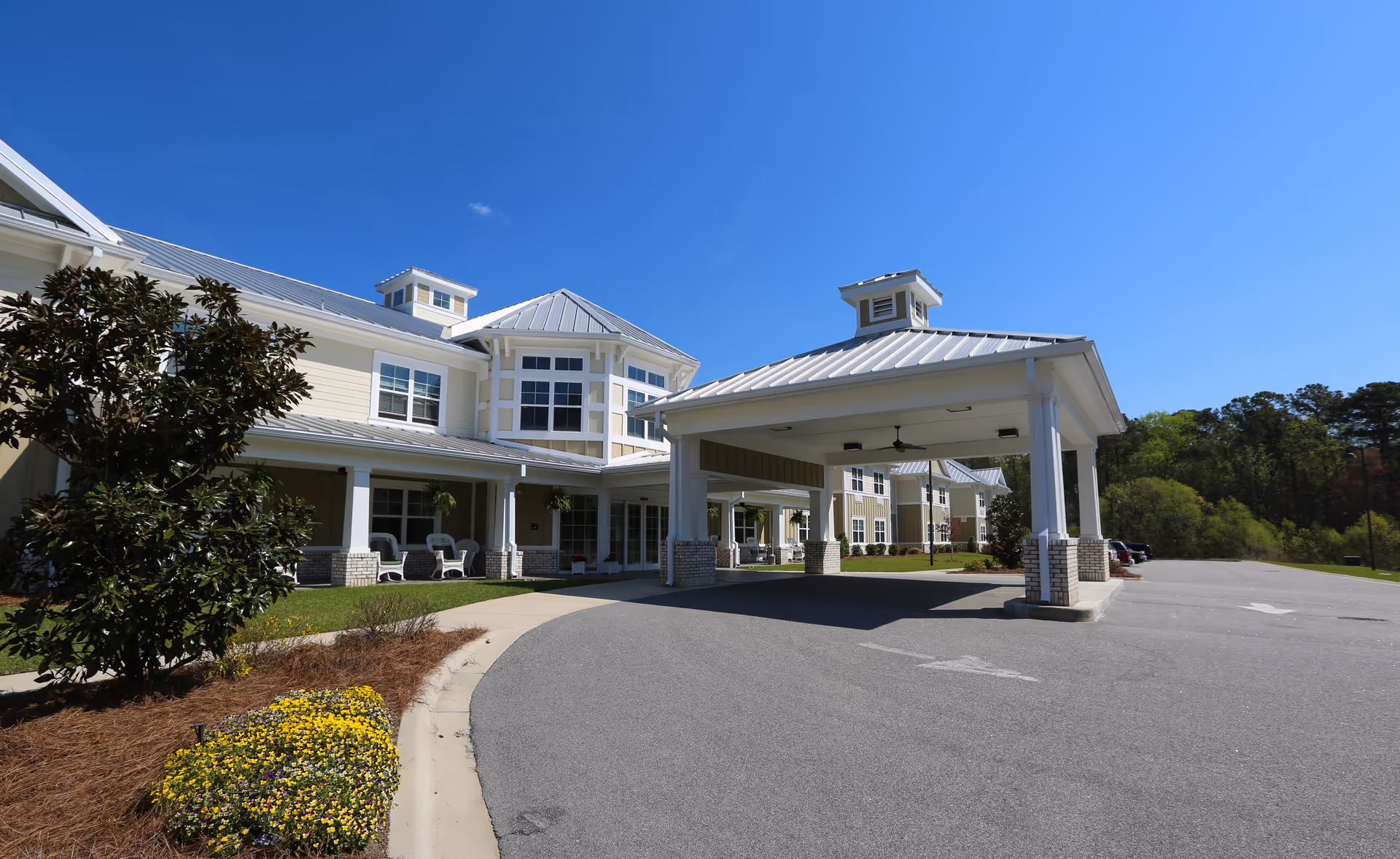 Exterior view of Arbor Landing at Ocean Isle senior living facility showing a large covered entrance with white pillars, a driveway, landscaped greenery including a tree and yellow flowers, and a clear blue sky.