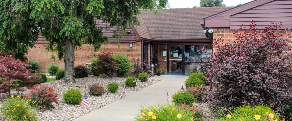 Exterior view of Rainelle Healthcare Center showing a brick building with a brown shingled roof. A concrete pathway leads to the glass entrance doors, flanked by landscaped garden beds with various shrubs, small trees, and American flags. The sky is partly cloudy.