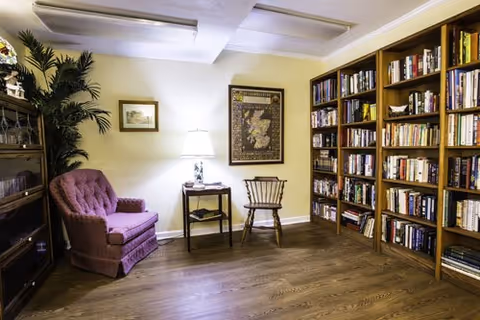 Cozy reading room with tall bookshelves, a pink upholstered armchair, a wooden chair and side table with a lamp on a hardwood floor.