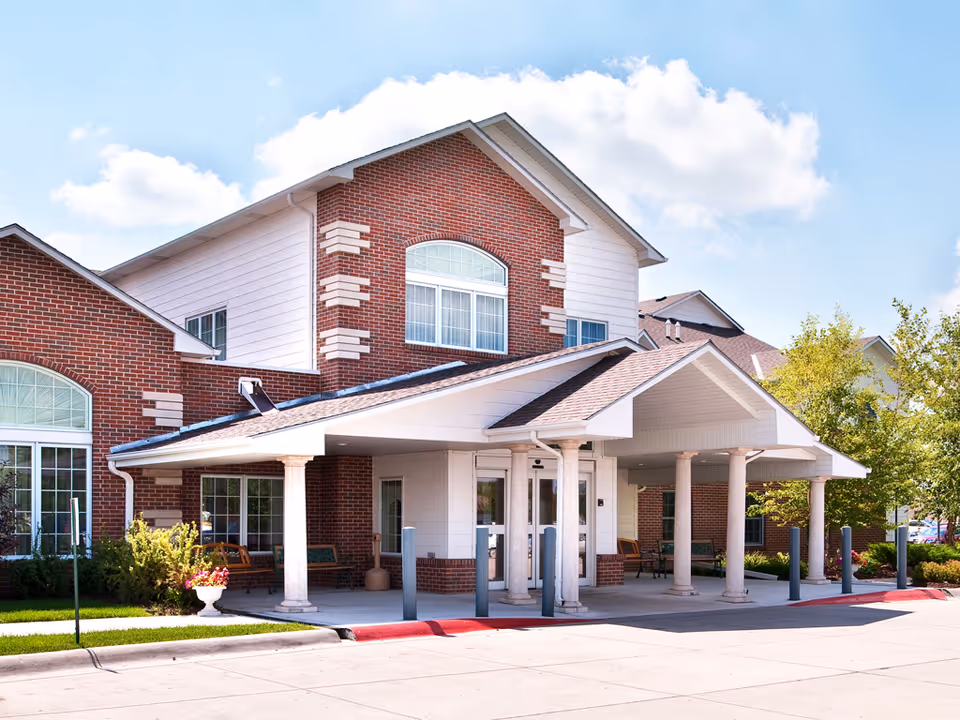 Exterior front view of a senior living facility building with red brick and white siding, featuring a covered entrance supported by columns, benches near the entrance, and landscaped greenery around the building under a partly cloudy sky.