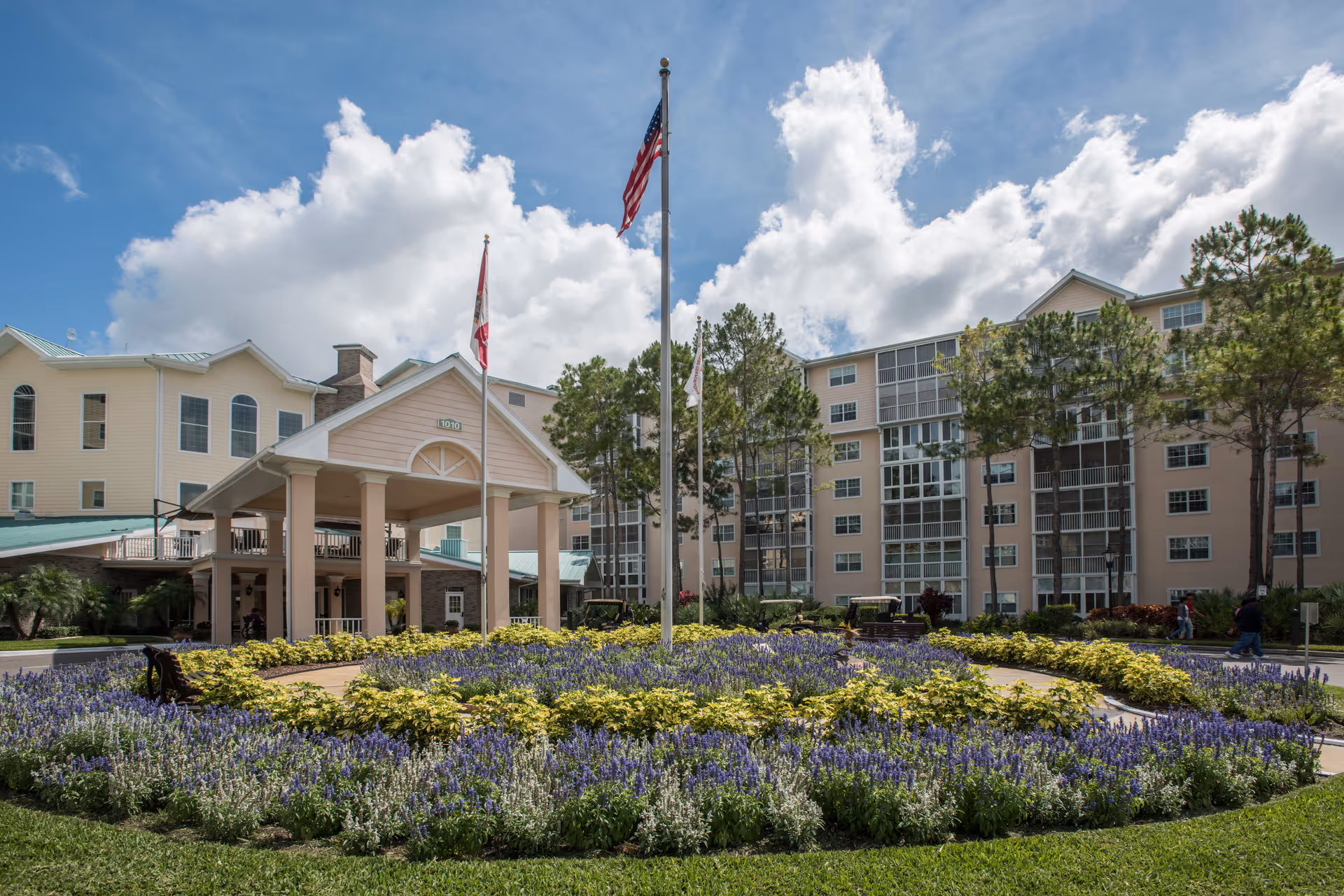 Front entrance of Freedom Plaza senior living facility with circular flower beds and flags.