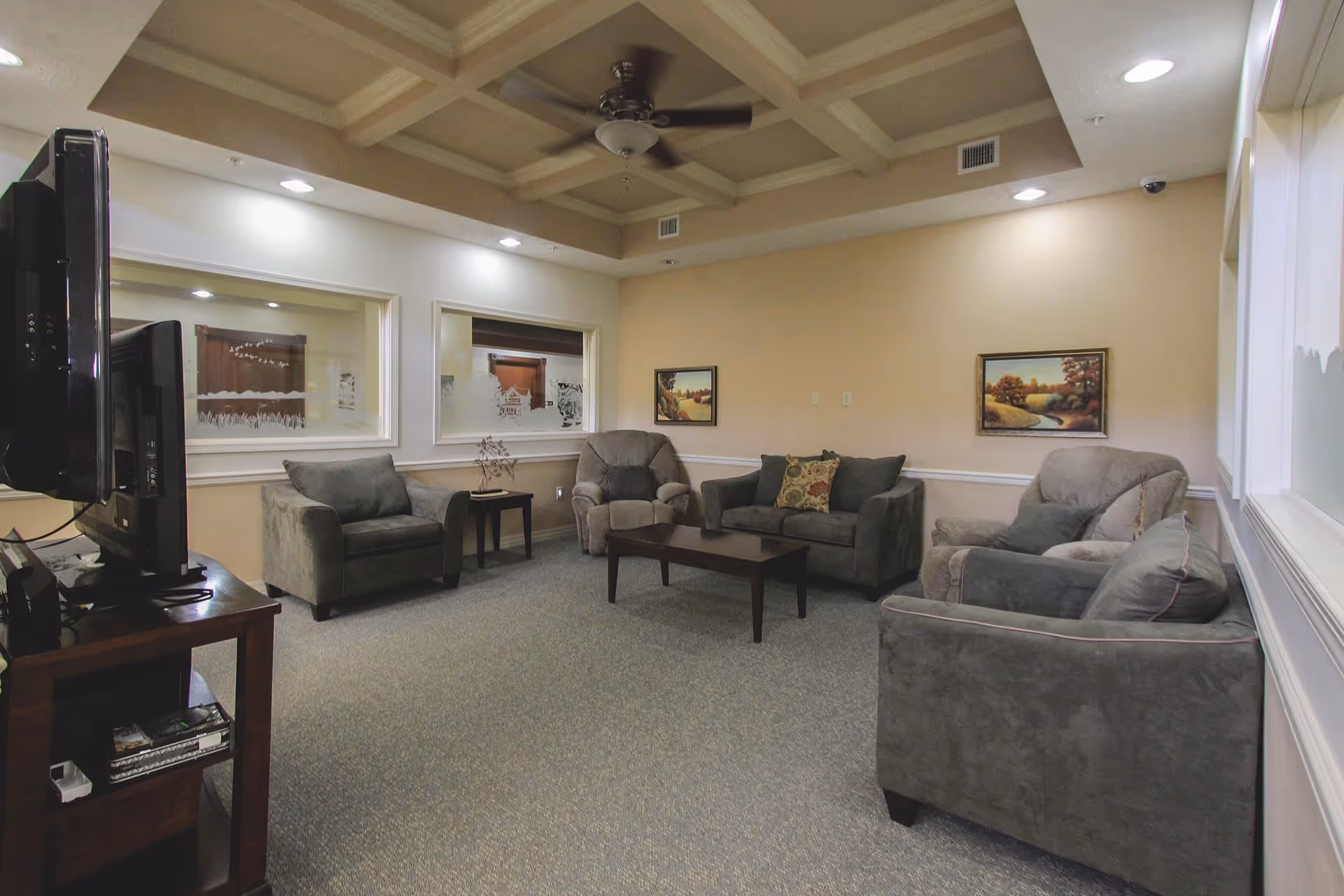 A cozy living room with gray upholstered armchairs and a loveseat arranged around a wooden coffee table. The room has beige walls with two landscape paintings, a coffered ceiling with a ceiling fan, recessed lighting, and windows with frosted glass looking into another room. A TV is positioned on a wooden stand to the left.