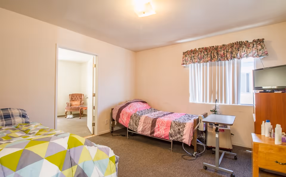 A simple bedroom with two beds, one with a colorful geometric patterned bedspread and the other with a pink and brown patterned bedspread. There is a window with vertical blinds and a floral valance, a small TV on a wooden dresser, and a bedside table with various bottles. An open door leads to another room with a pink armchair and a small table.