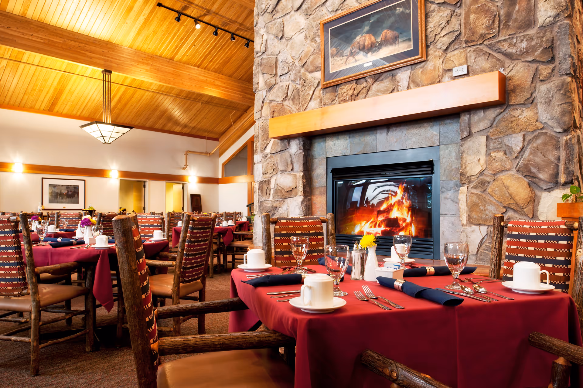 Dining room with tables covered in red tablecloths, set with white cups, glasses, silverware, and napkins. The room features wooden chairs with patterned upholstery, a stone fireplace with a fire burning, and a wooden ceiling with warm lighting.