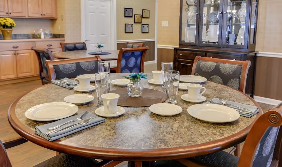Round dining table set for six with plates, cups, glasses and napkins in a communal dining room.