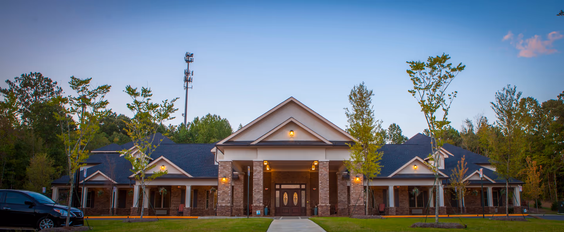 Front exterior view of Magnolia Senior Living at Sugar Hill building with a brick facade, large entrance with double doors, and a covered porch supported by columns. There are young trees and a parked black car in front, with a clear sky in the background.