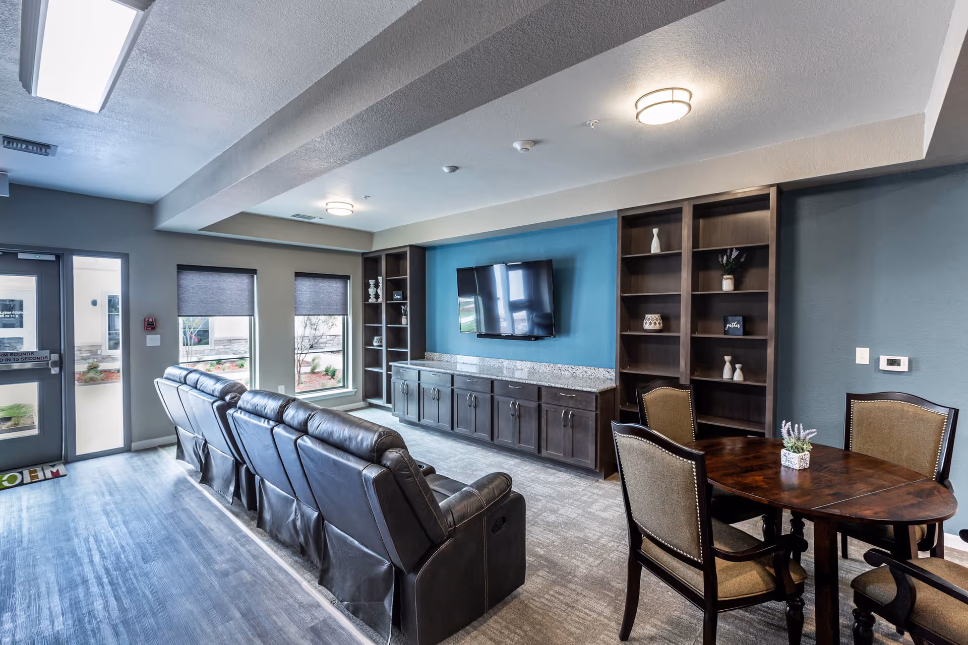 A modern living room area with a row of black leather recliners facing a wall-mounted flat-screen TV. The wall behind the TV is painted blue and flanked by dark wood built-in shelves and cabinets. To the right, there is a round wooden table with four upholstered chairs. The room has large windows with gray blinds and a door leading outside. The floor is a combination of wood and carpet.