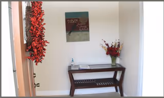 Entryway area with a wooden bench and a vase of flowers on top. A decorative wreath with red berries hangs on the left side of the door, and a framed wall art piece is mounted above the bench. A bottle of hand sanitizer and some papers are placed on the bench.
