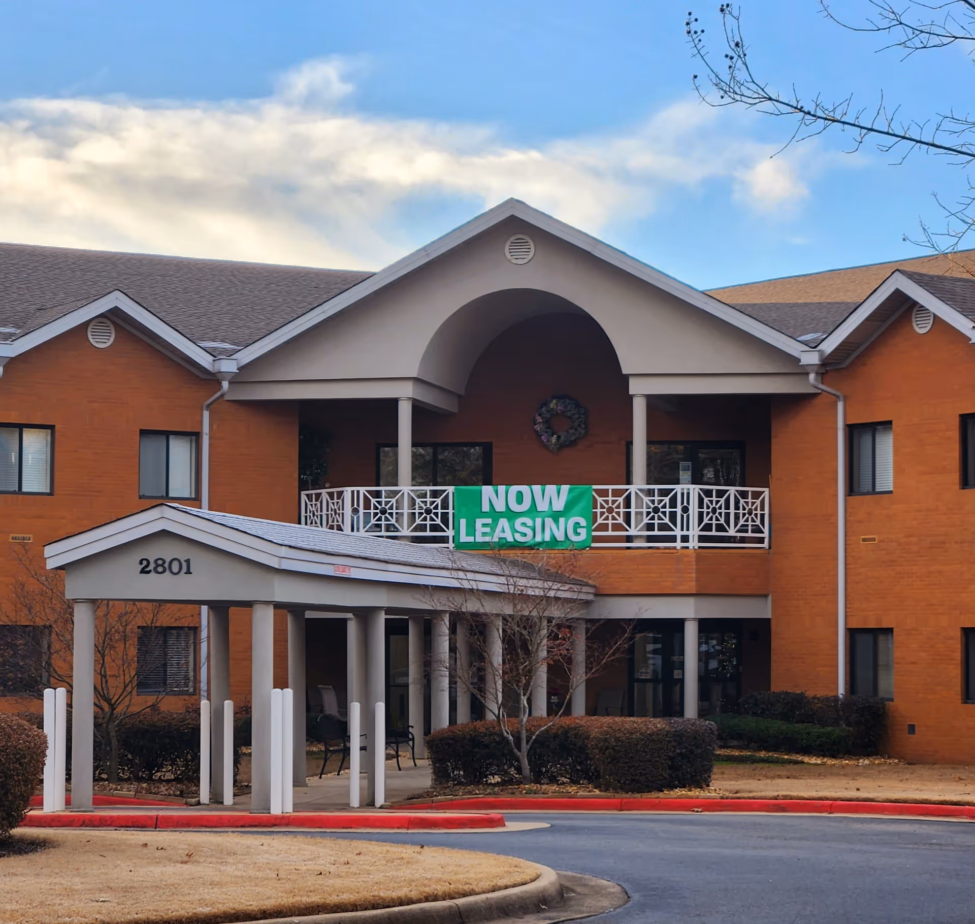 Exterior view of a two-story brick building with a covered entrance and a balcony displaying a green banner that reads 'NOW LEASING'. The building number 2801 is visible on the entrance structure. The sky is partly cloudy and there are some leafless trees and bushes around the building.