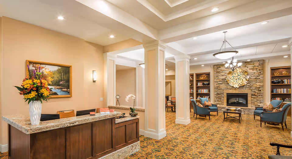 Reception area with a granite countertop desk adorned with a vase of colorful flowers and a painting on the wall. The space opens into a cozy lounge area featuring a stone fireplace, built-in bookshelves, and several blue upholstered chairs arranged around a coffee table. The room has warm lighting and a patterned carpet.