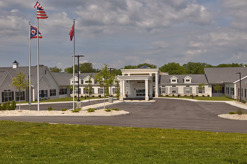 Front exterior view of Traditions of Lebanon facility showing a large single-story building with a covered entrance, multiple windows, and three flagpoles flying the American flag, Ohio state flag, and another flag. The building is surrounded by a paved driveway, landscaped areas with small trees and shrubs, and a grassy lawn under a partly cloudy sky.