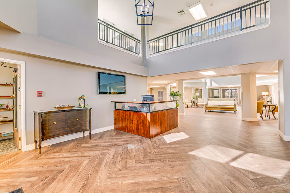 Bright two-story senior living lobby with a reception desk, seating area, and herringbone wood flooring.