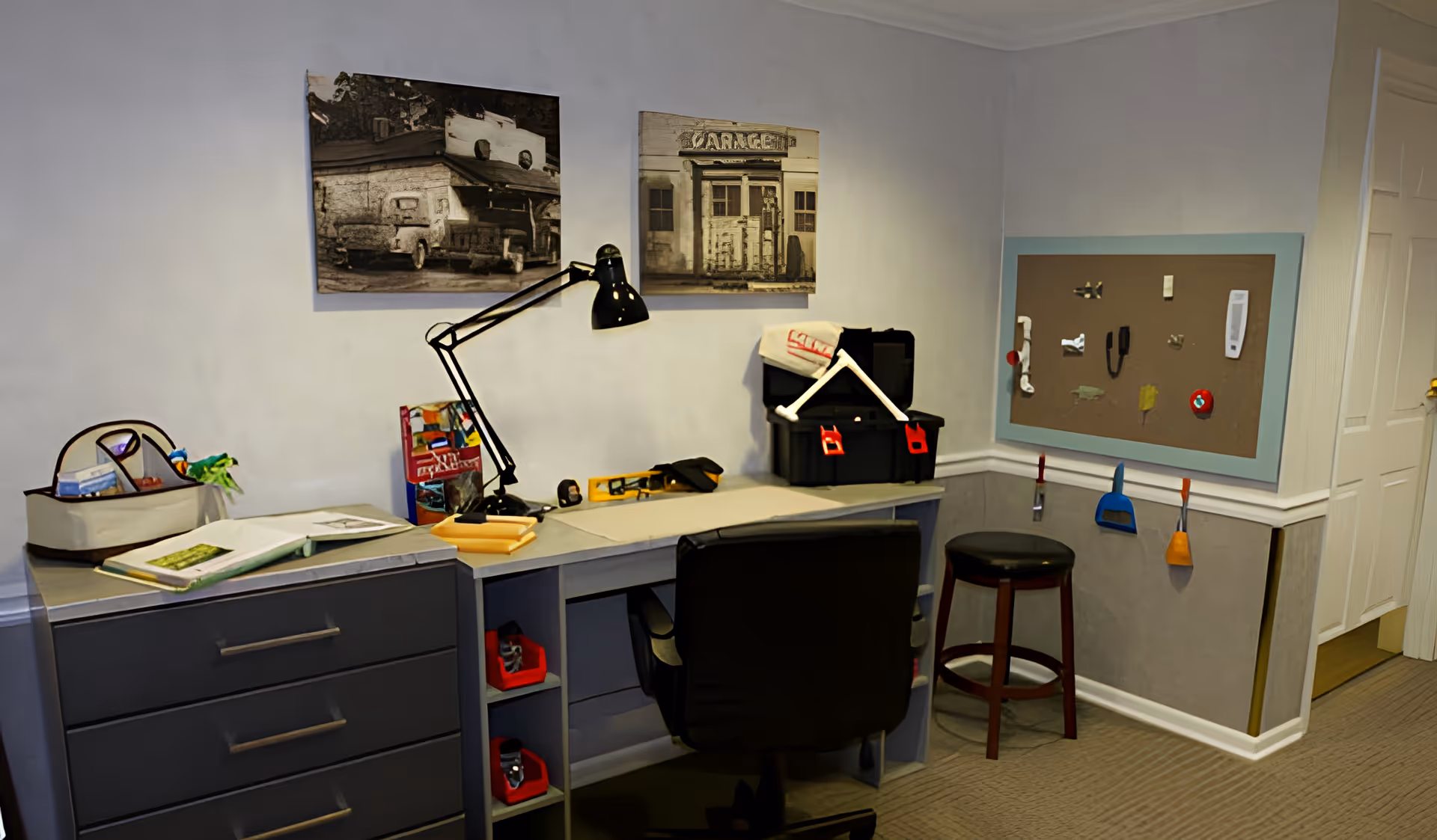 A small workspace area with a corner desk, black office chair, and a black desk lamp. On the desk are various items including a toolbox, books, and small tools. Above the desk are two black and white photos of old buildings. On the wall to the right is a bulletin board with assorted small items pinned to it, and below it hang small cleaning tools. The room has light-colored walls and carpeted flooring.