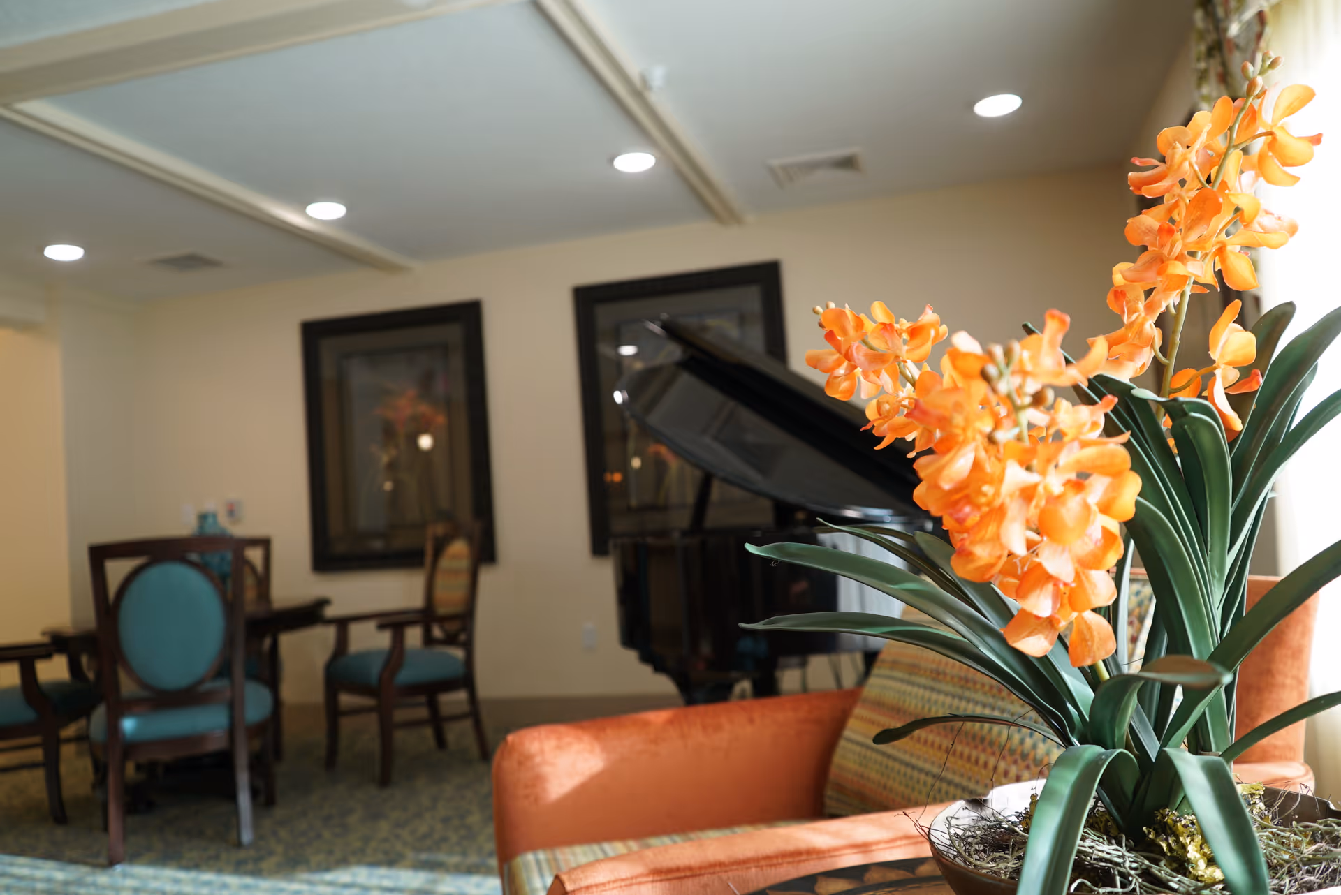 A cozy interior room featuring a bright orange flower arrangement in the foreground on a table, an orange armchair, a grand piano in the background, and a dining table with teal upholstered chairs. The walls are decorated with framed artwork.