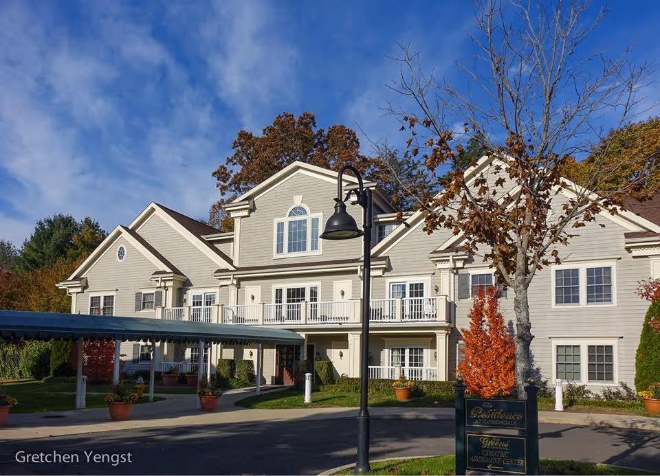 Front exterior of a two-story senior living building with balconies, a covered drop-off, a lamp post, and autumn trees.
