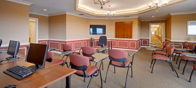 A classroom-style meeting room with multiple desks and chairs arranged facing a wall-mounted TV. The room has beige walls with white trim, a carpeted floor, and a ceiling with recessed lighting and decorative molding. There are computer monitors on desks in the foreground.