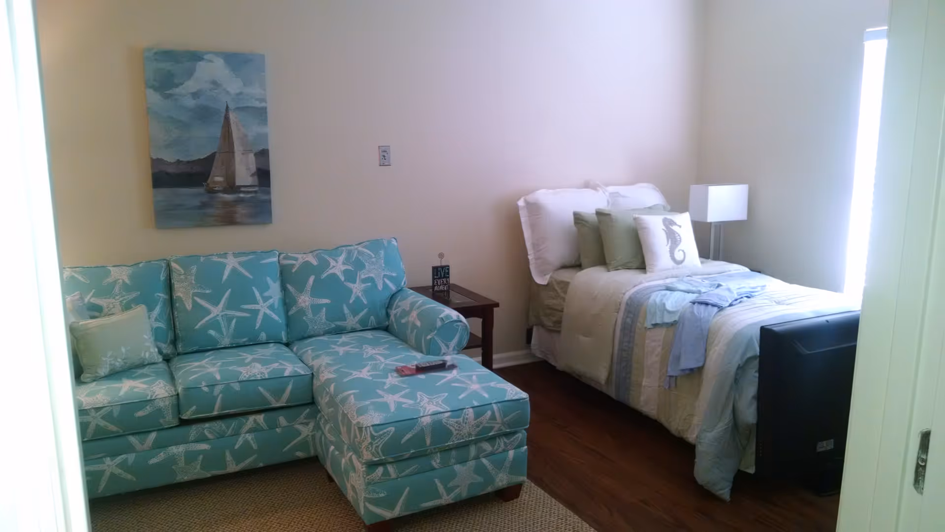 A small bedroom with a single bed made with white and light blue bedding, several pillows including one with a seahorse design, a turquoise couch with white starfish patterns, a wooden side table with a small decorative sign, a white lamp, and a painting of a sailboat on the wall.