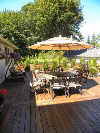 Outdoor wooden deck area with a large patio table surrounded by six ornate metal chairs with cushions. A large beige umbrella provides shade over the table. There is a swing bench on the left side and potted plants along the railing. Trees and greenery are visible in the background under a clear blue sky.