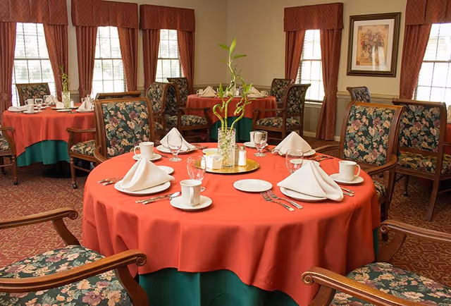 Dining room with round tables dressed in red tablecloths, floral-upholstered chairs, place settings, and a small plant centerpiece.