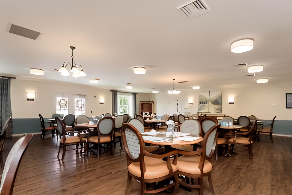 A spacious dining room with multiple round wooden tables and cushioned chairs arranged neatly. The room has wooden flooring, soft lighting from ceiling fixtures, and windows with curtains allowing natural light to enter. The walls are painted white with a blue lower half, and there is artwork hanging on the walls.