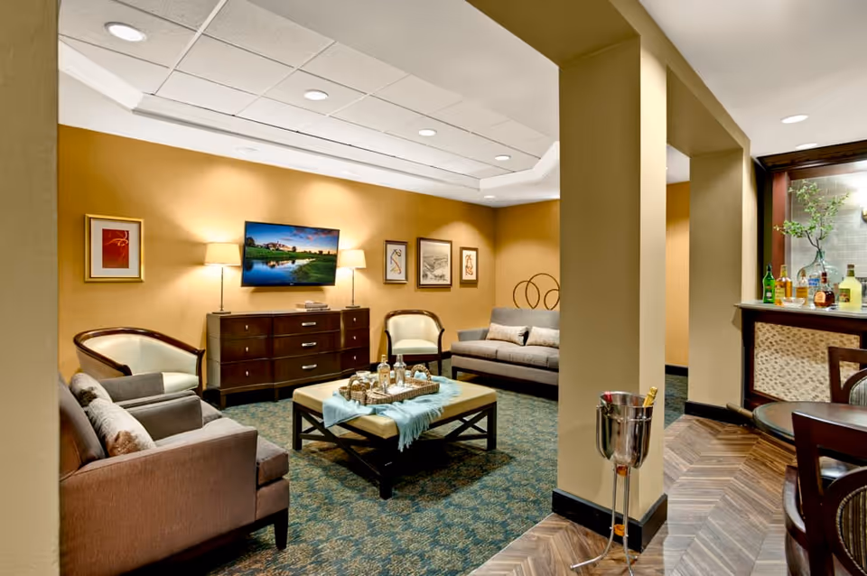 A cozy living room area with beige walls and carpeted floor. The room features two sofas and two armchairs arranged around a central ottoman table with a tray holding bottles and glasses. A flat-screen TV is mounted on the wall above a dark wooden dresser, flanked by two table lamps. There are framed artworks on the walls. To the right, there is a small bar area with bottles and glasses, and a round table with chairs. The room is softly lit with recessed ceiling lights.